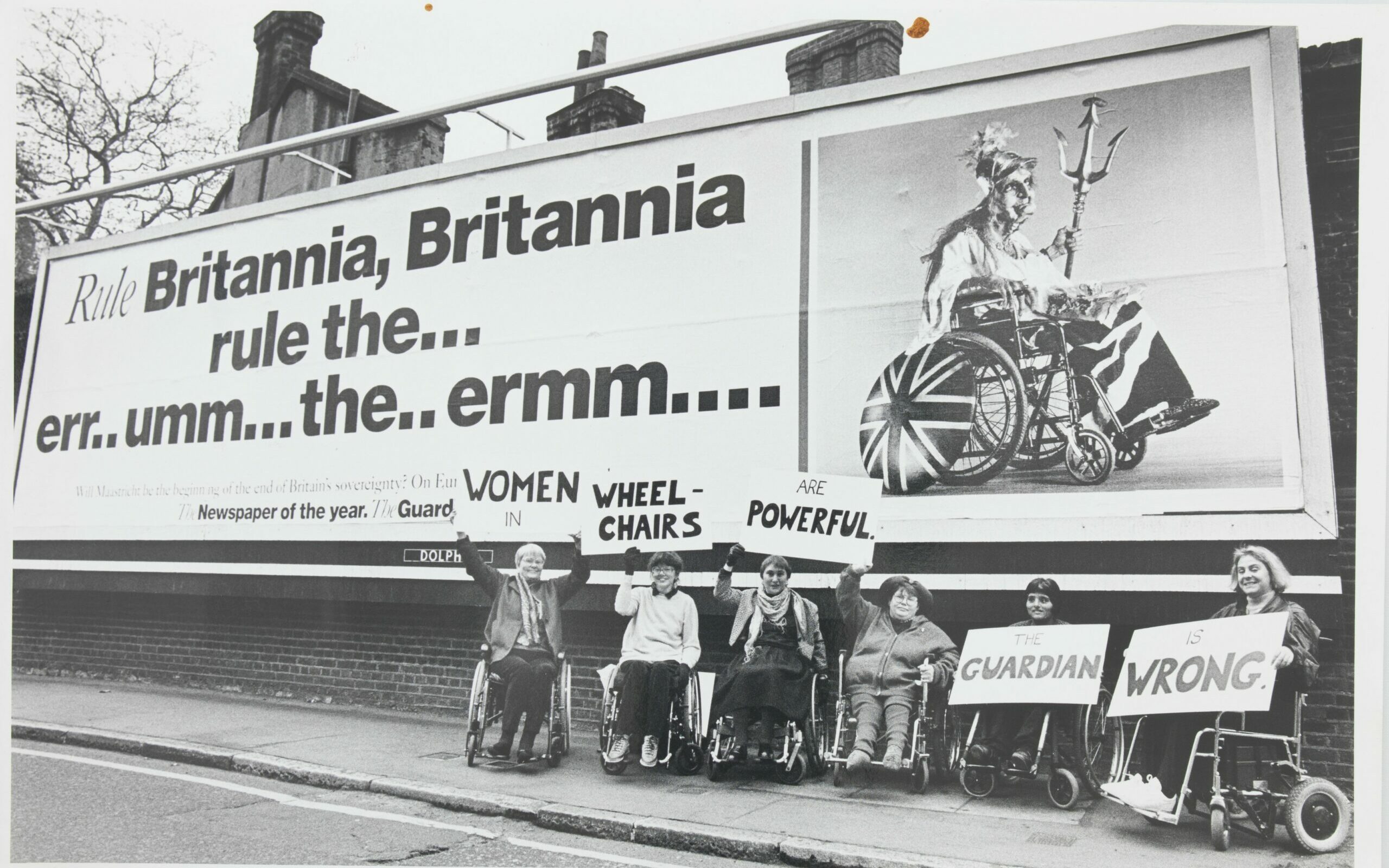 A black and white photograph of six disabled women gathered beneath an advertising billboard. The women carry signs which read ‘women in wheelchairs are powerful the Guardian is wrong’.
