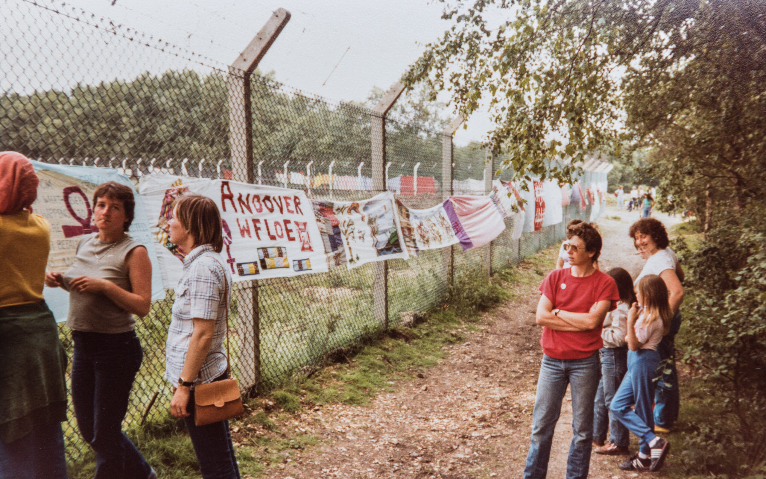 A colour photograph of five women and two young girls standing casually along a dirt track which runs next to a tall chain link fence. A long line of colourful handmade banners are decorating the fence. One woman looks directly at the camera whilst everyone else is admiring the banners.