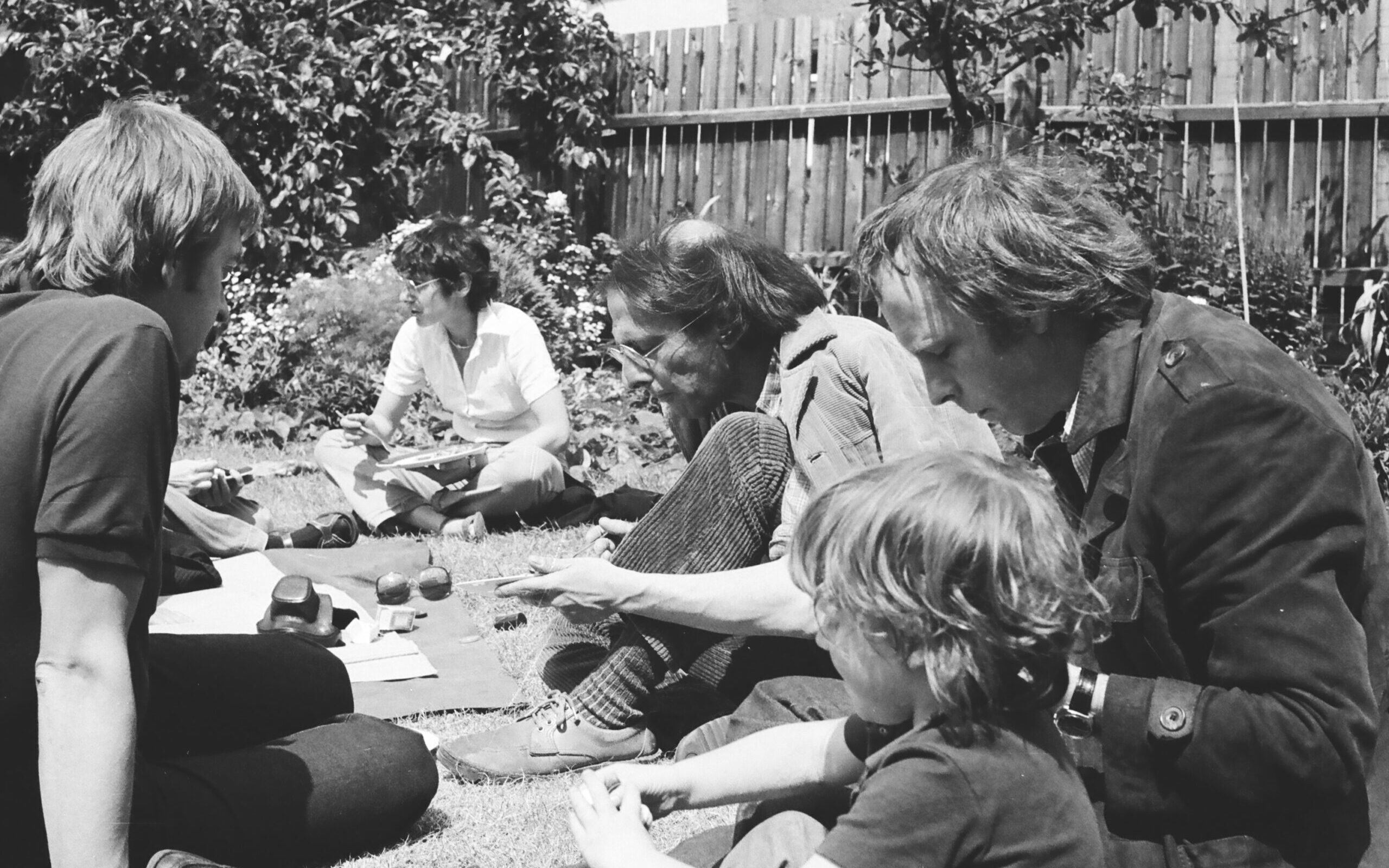 A close up black and white photograph showing three white men, one white woman and one young white boy sitting together on the grass in someone’s garden. They are having a picnic and seem deep in conversation. There is a hedge and a wooden fence in the background.