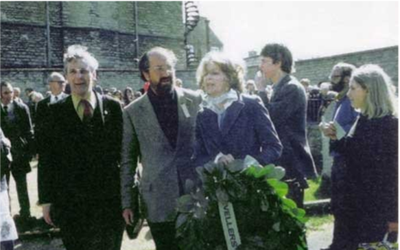 A grainy colour photograph of a man and woman, Benn and his wife Caroline, standing in a crowded and busy area. They are dressed smartly, and the woman is carrying a leafy commemoration posy with a white ribbon across it that reads 'Levellers'.