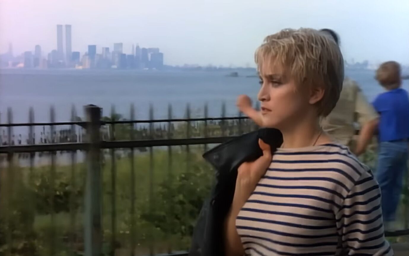 A screen shot from a 1980s colour music video featuring Madonna. A young white woman with cropped blonde hair, wearing a striped top is walking along a river path looking lost in thought. The New York skyline is visible in the background.