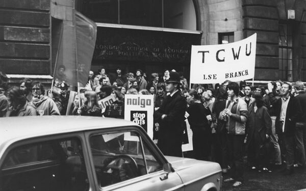 Protestors gathered outside an LSE building, holding placards including for the TGWU union. A police officer stands by.