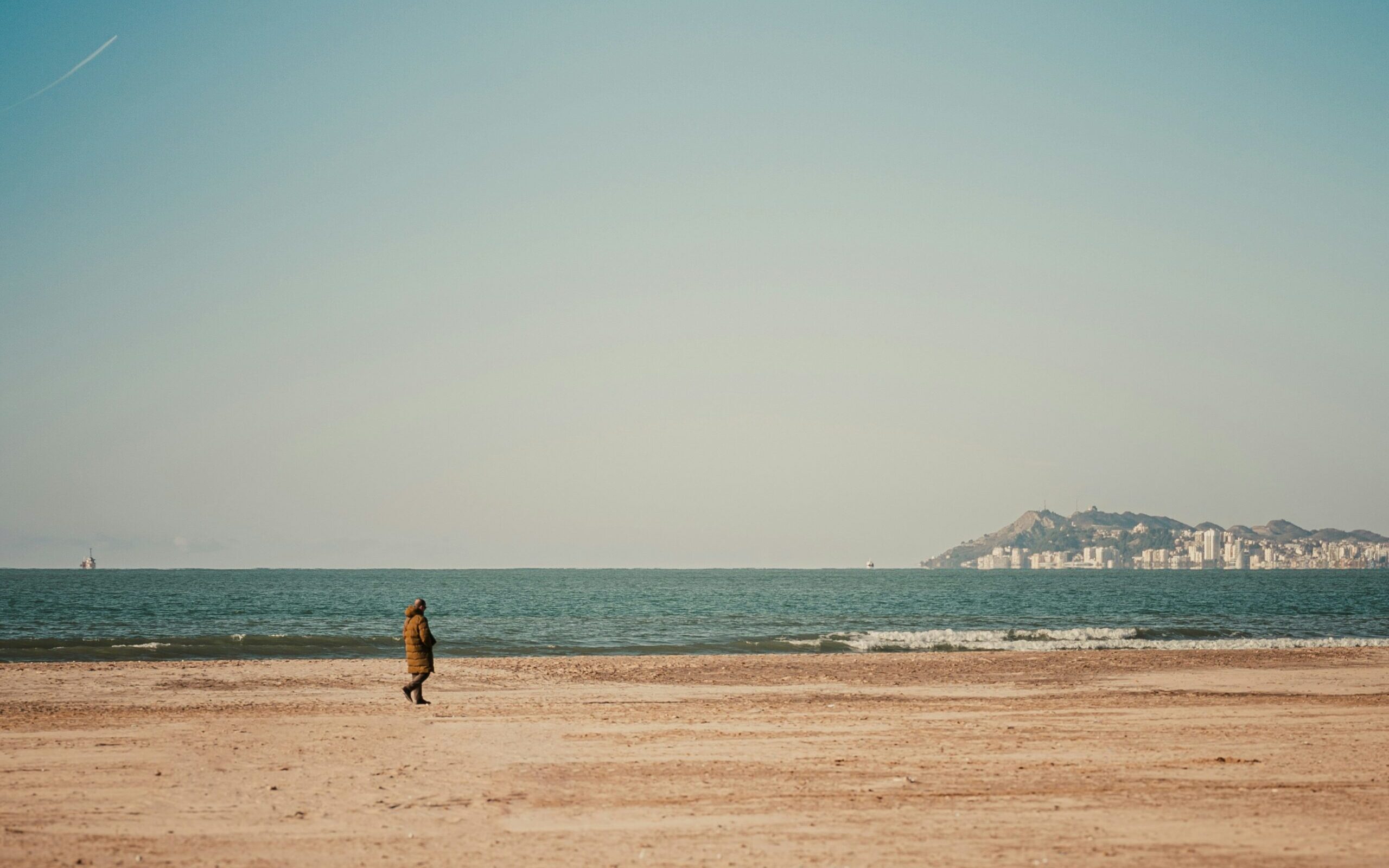 A colour photograph depicting a long figure walking along a deserted sandy beach.