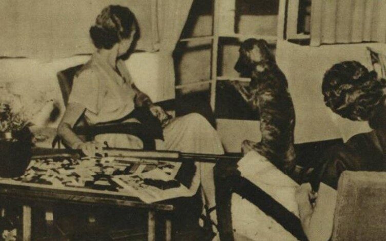 A sepia photograph showing two white women and a dog in a comfortable living room in central Kenya. The dog is looking to the shuttered window as if it has heard something, and one woman is reaching for a large rifle to her side.
