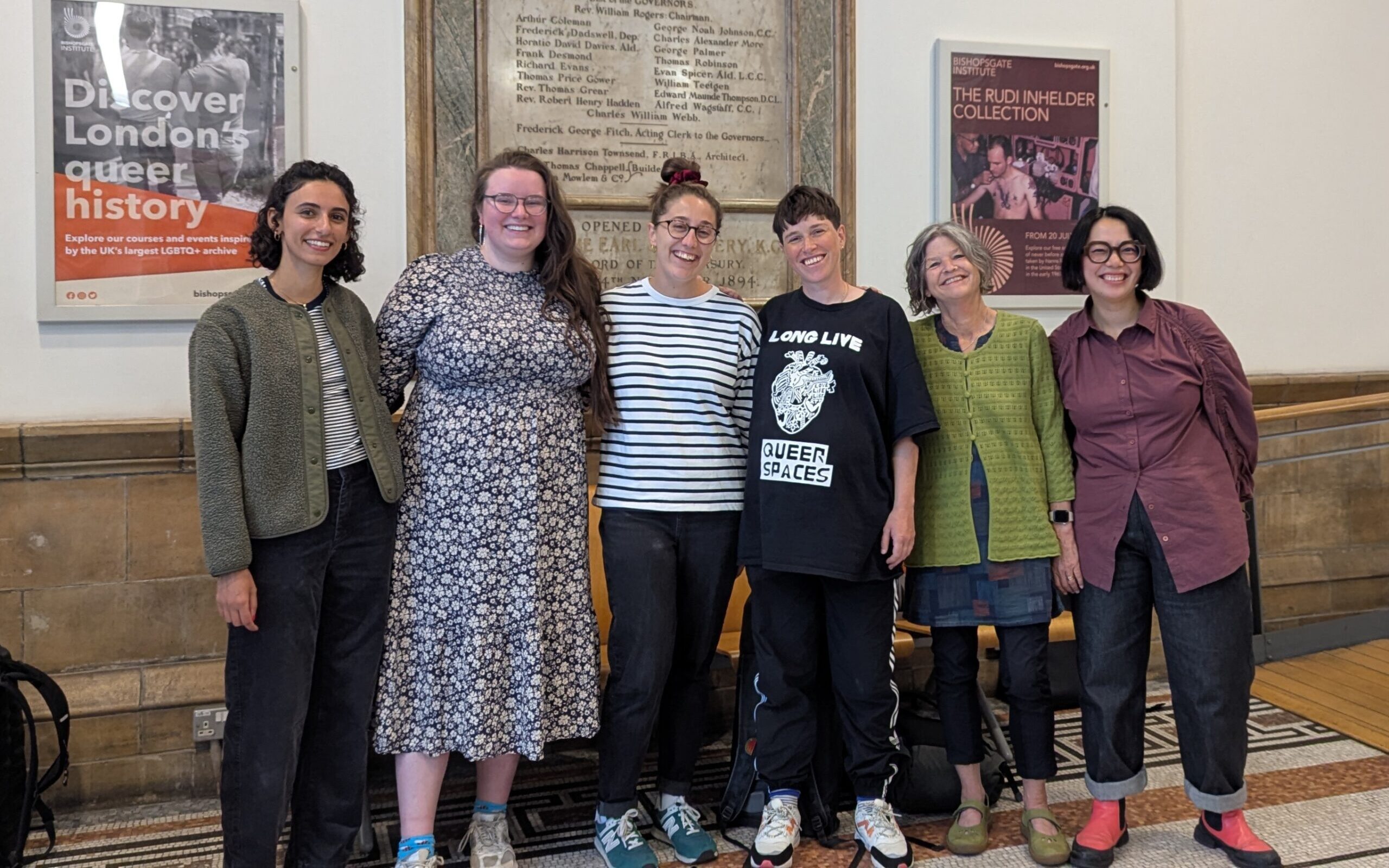 A colour landscape photograph of six people stood closely together in a collegial way. They are all smiling broadly at the camera. Behind them is a wall with two events posters and a large foundation stone.