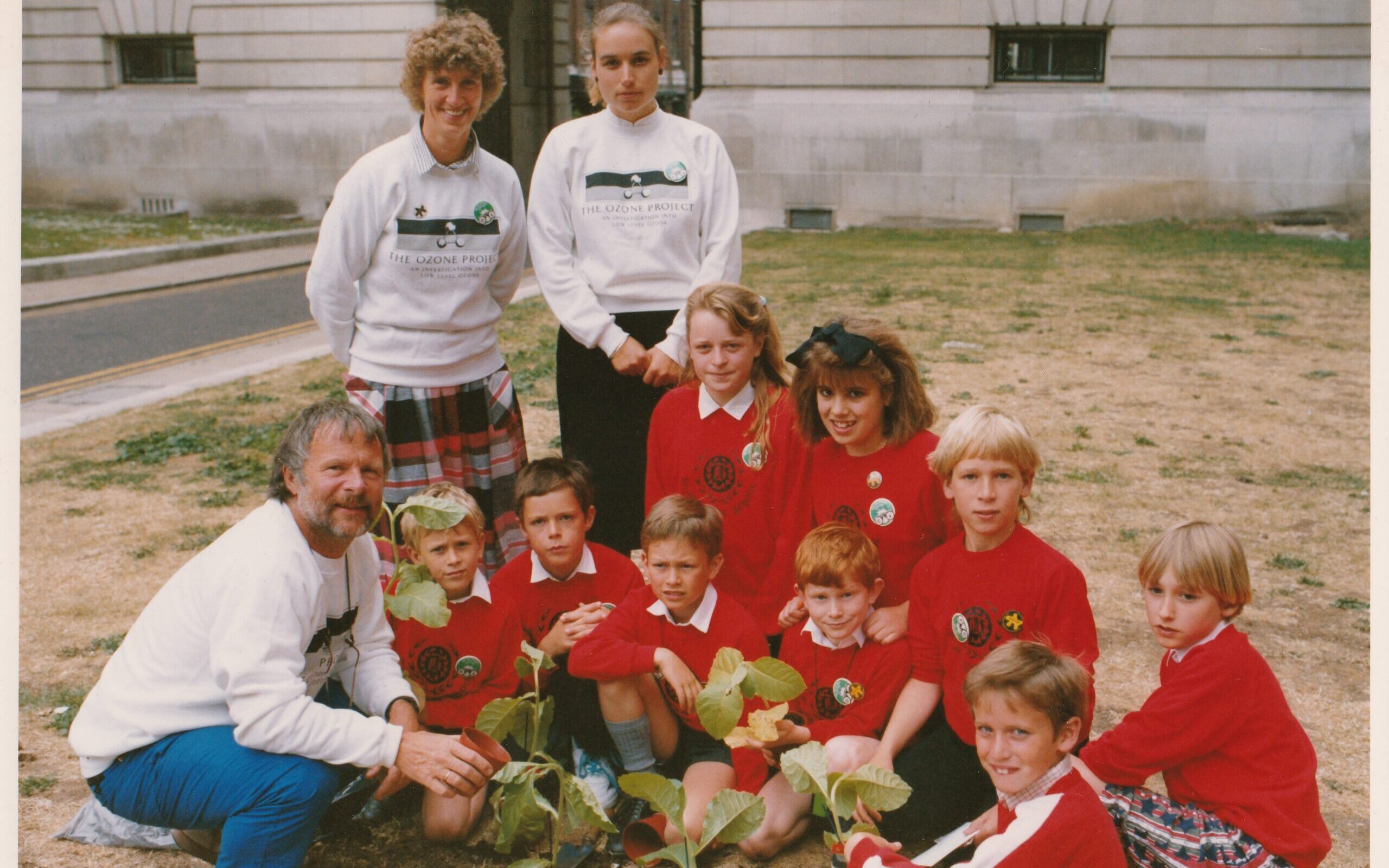 3 adults in The Ozone Project t-shirts pose with 9 children in matching red sweatshirts. In front of them are some plants they've just planted.