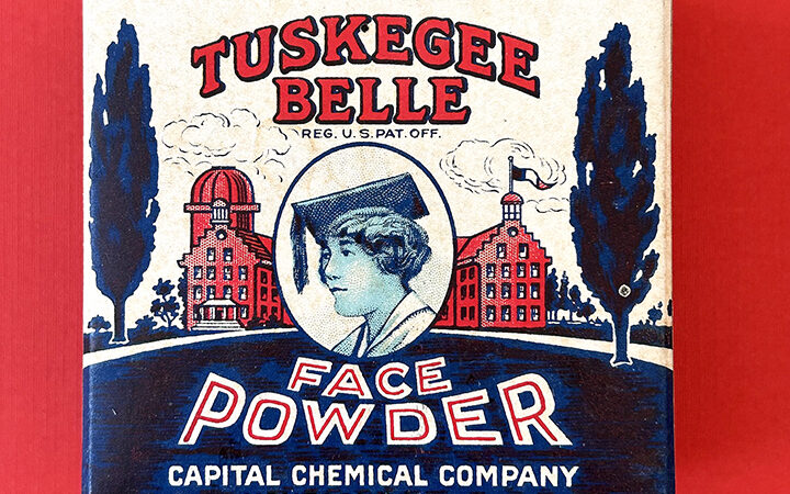 A 1920s Tuskegee Belle Face Powder box with a red, white and blue theme. In the centre of the box is the profile of a young Black woman, wearing an academic mortar board and gown. In the background are grand university buildings and a lawn and trees.