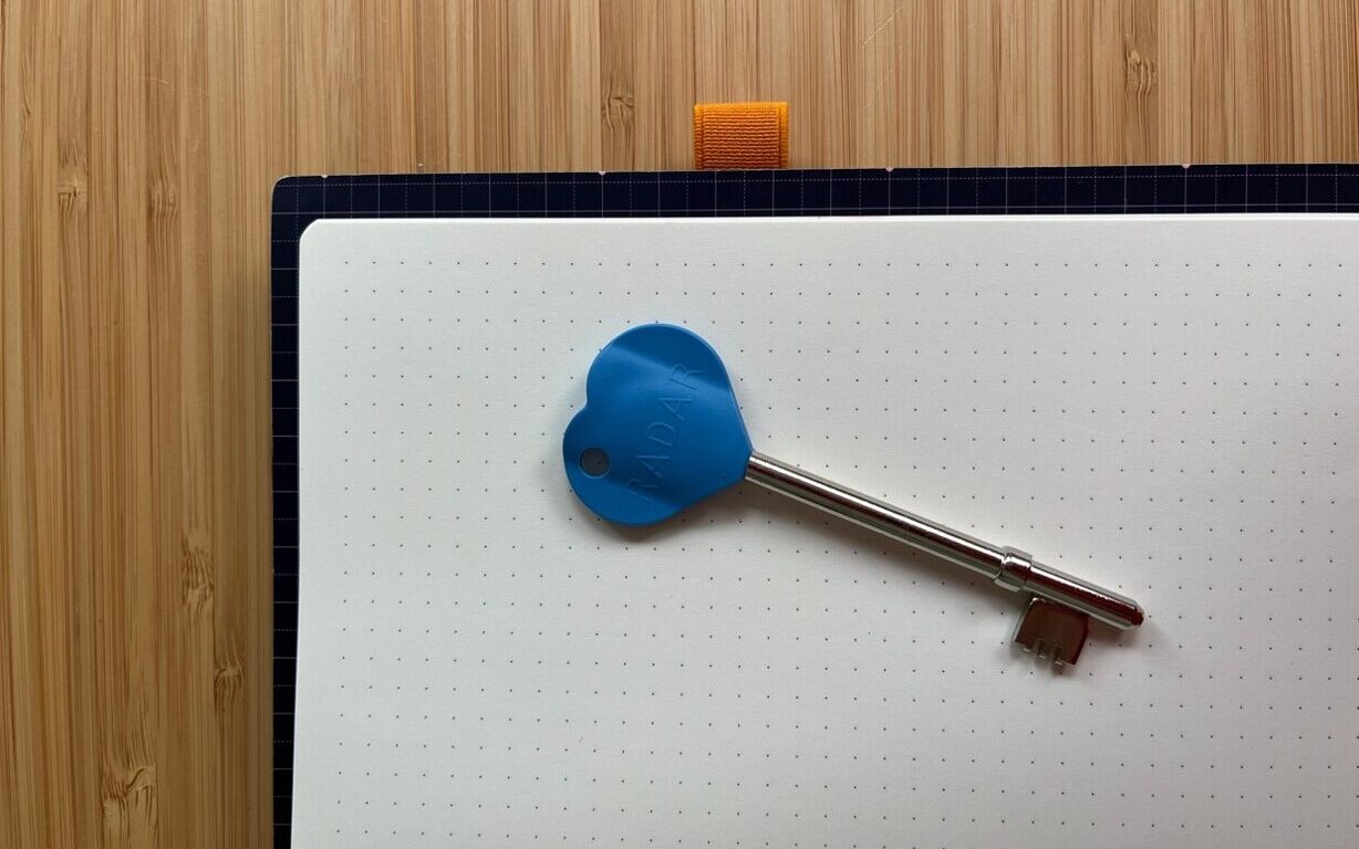 Radar Key with a blue heart-shaped top and a three-pronged key, rested on a dotted notebook on a light brown, wooden table.