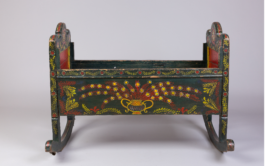 A dark green wooden cradle on curved rockers, with arched headboards at each end. The long side is painted with a two-handled yellow vase holding sprays of red leaves and yellow flowers, framed by yellow and red foliage. A band of trailing vines and small red flowers runs along the top edge; the inner end panels are red. The paint is worn and chipped in places.