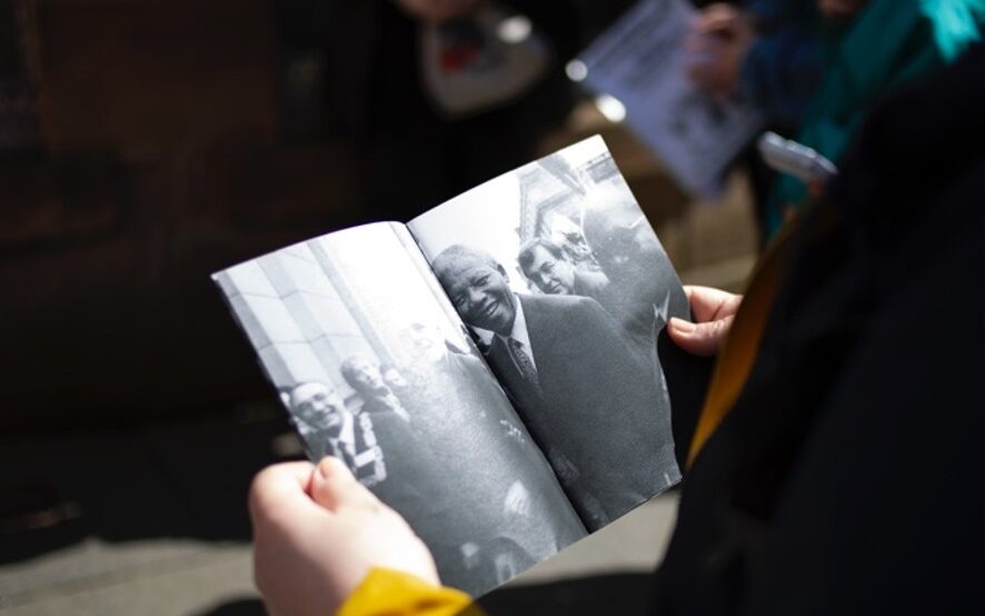 21st century photograph of a person stood outside holding open a pamphlet. In the pamphlet is a photo of Nelson Mandela. In the background, someone else is reading another pamphlet. They are outside.